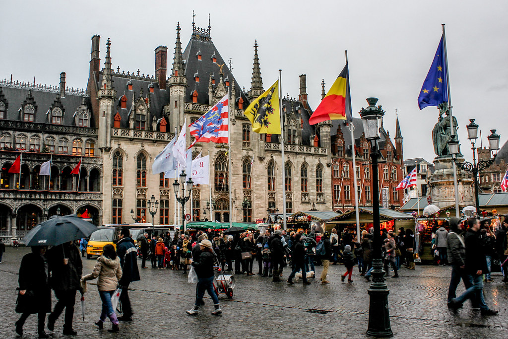 Ambiente animado en la plaza Markt de Brujas con la fachada gótica del edificio histórico y banderas belgas y europeas.