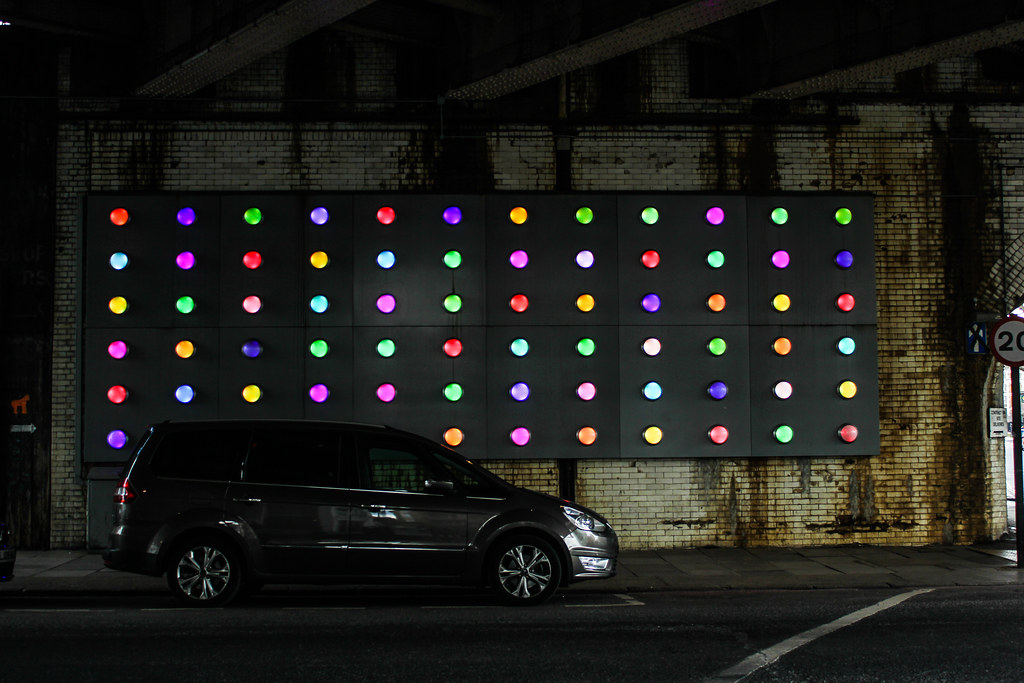 Panel de arte urbano iluminado con luces de colores y coche estacionado.