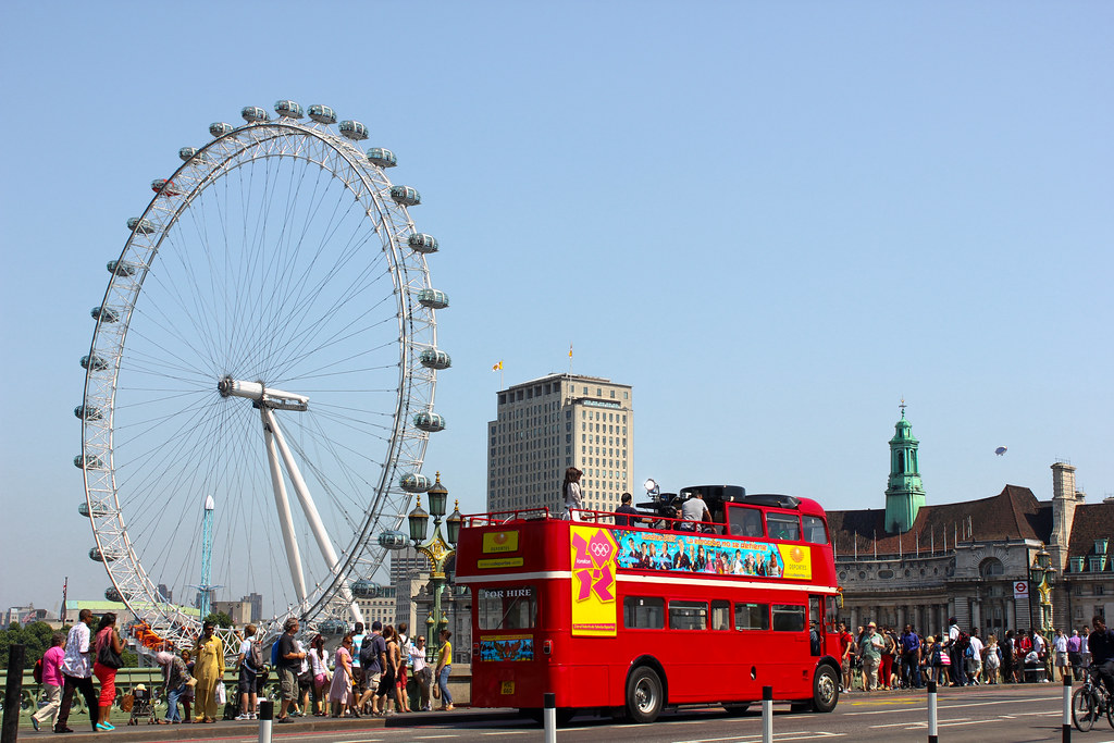 Autobús turístico rojo de dos pisos pasando por el London Eye lleno de visitantes.