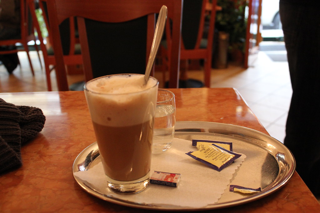 Café latte en vaso transparente con cuchara y azúcar sobre bandeja metálica en cafetería acogedora.