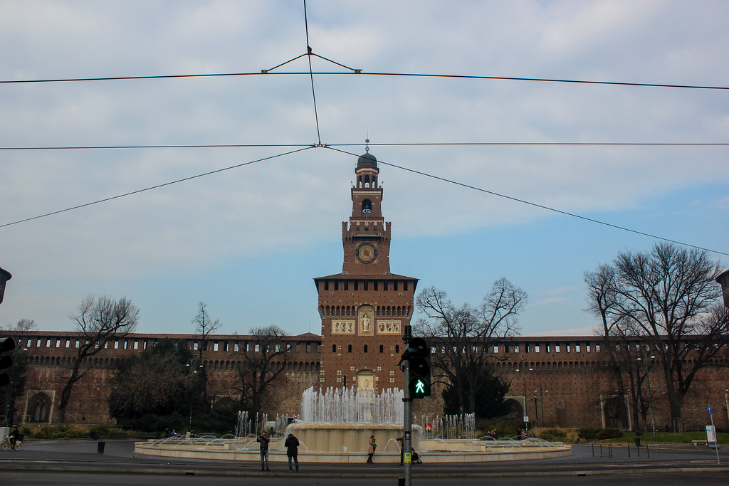Castillo Sforzesco en Milán visto desde la calle con una fuente en primer plano y cables de tranvía cruzando el cielo.