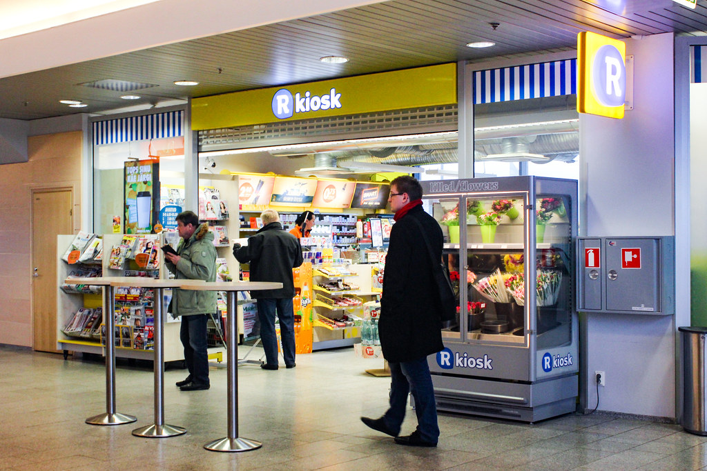 Clientes en un R-kiosk en el interior de una estación con revistas y aperitivos.
