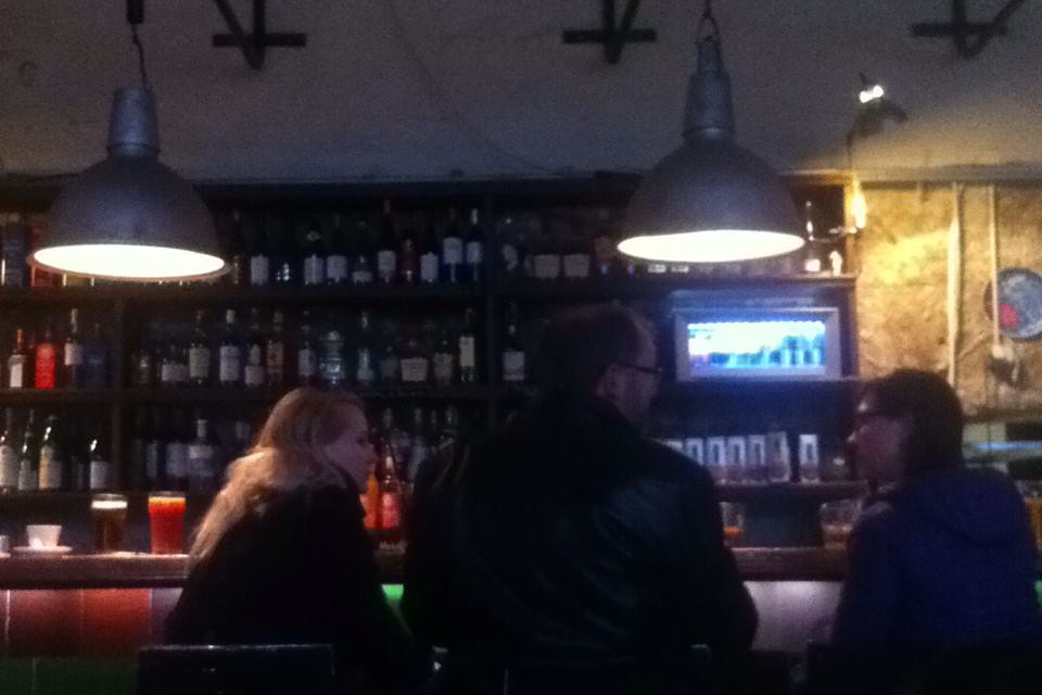 Clientes sentados en la barra del restaurante F-Hoone con iluminación ambiental y estantes de botellas al fondo.