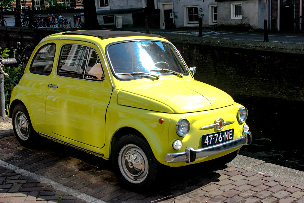 Coche clásico Fiat 500 amarillo aparcado junto a un canal en una calle adoquinada de Ámsterdam.