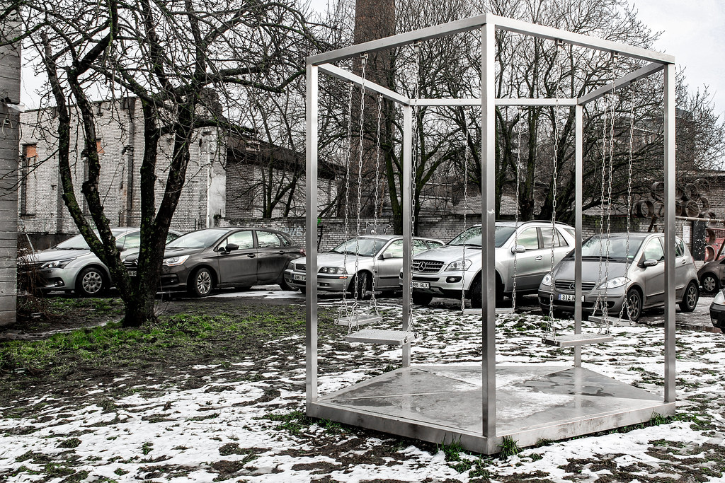 Columpio metálico moderno en un aparcamiento con nieve y árboles desnudos.