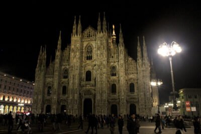 Catedral de Milán iluminada por la noche con gente caminando en la plaza.