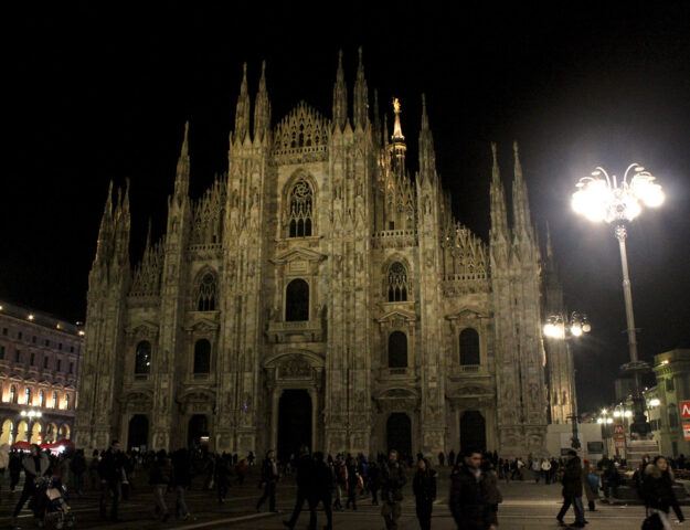 Catedral de Milán iluminada por la noche con gente caminando en la plaza.
