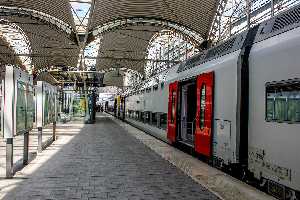 Estación de tren de Lovaina con tren de doble piso y techo de vidrio arqueado en un día soleado.
