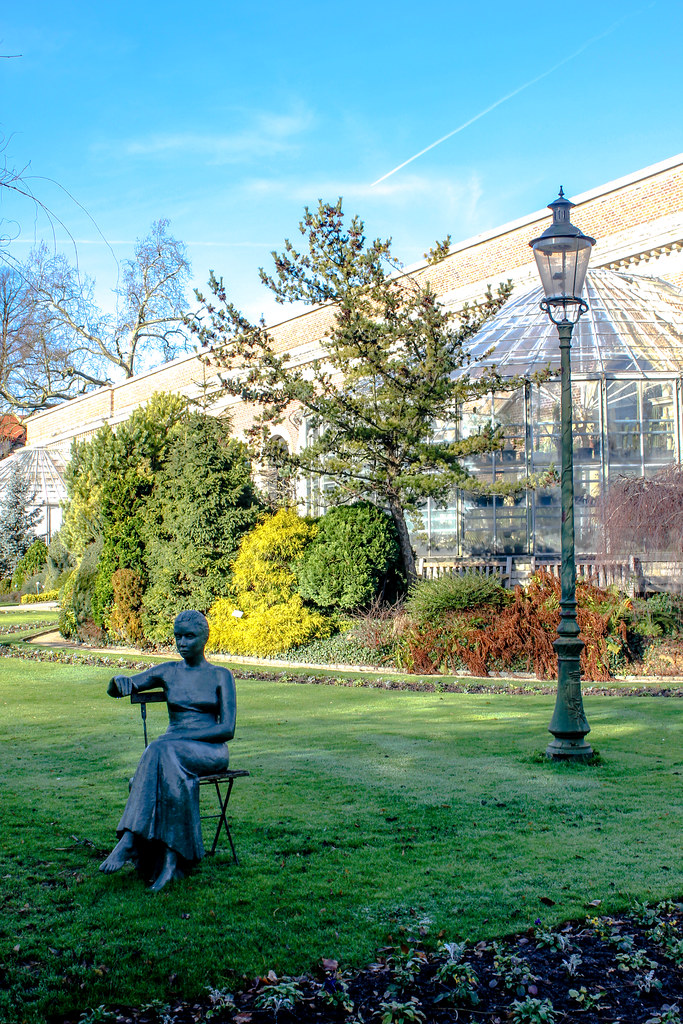 Estatua de mujer sentada en una silla en el Jardín Botánico de Lovaina con invernadero al fondo.
