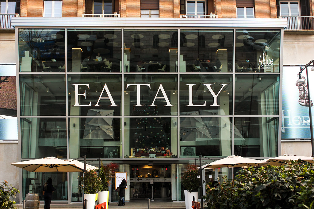 Fachada del mercado gastronómico Eataly en Milán con visitantes y sombrillas exteriores.