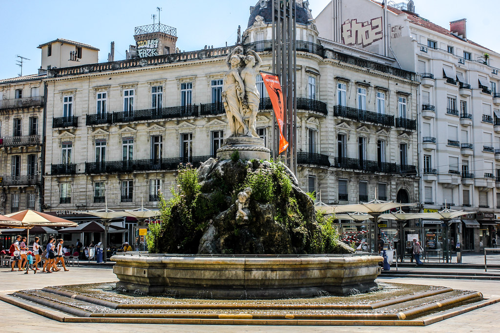 Fuente de las Tres Gracias en la Place de la Comédie, Montpellier, con edificios históricos al fondo.