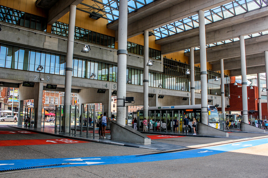 Interior abierto de la estación de autobuses de Lovaina con pasajeros y vehículos en espera.