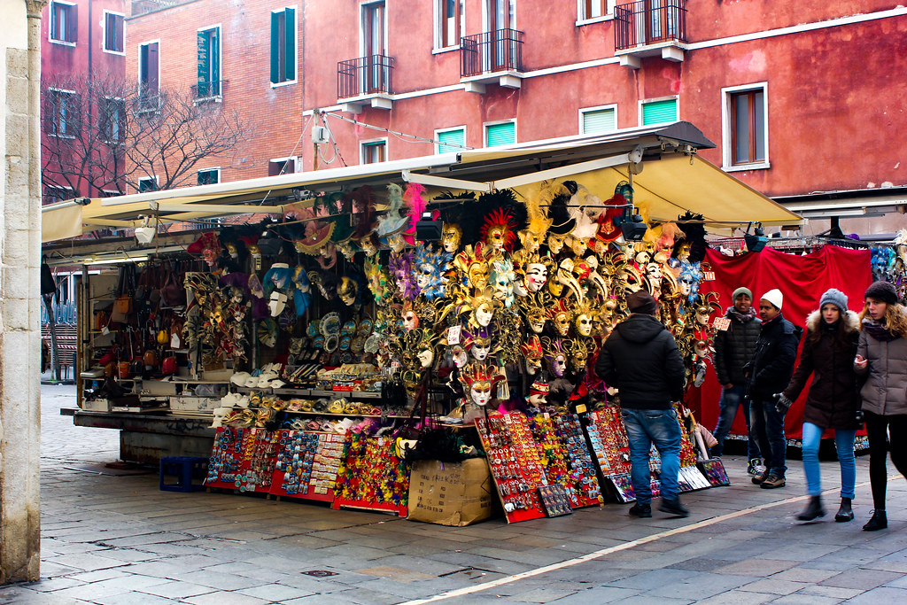 Puesto de venta de máscaras venecianas y souvenirs con transeúntes en una calle de Venecia.