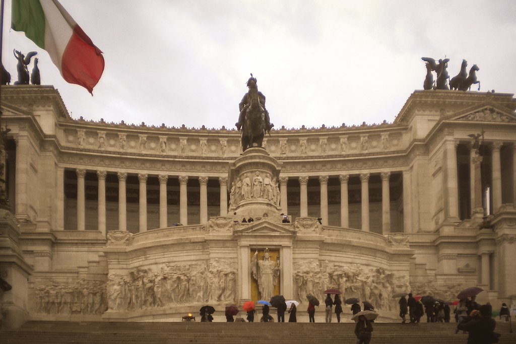 Monumento a Vittorio Emanuele II en Roma con visitantes en un día lluvioso y bandera italiana ondeando.