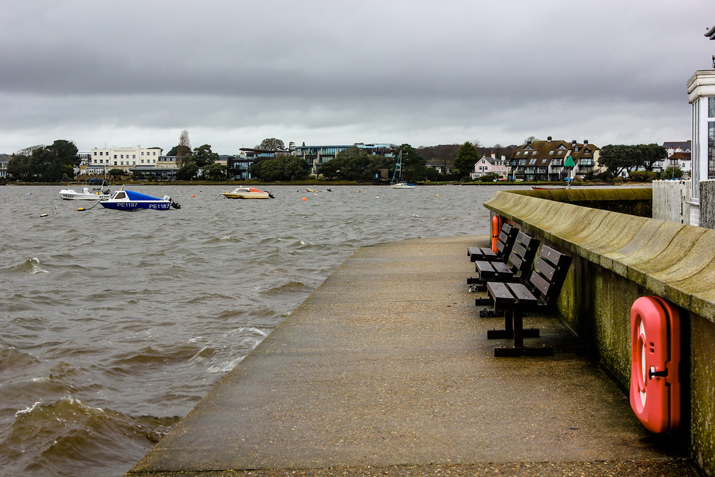 Muelle de Mudeford Quay con bancos vacíos y botes flotando en un día gris.