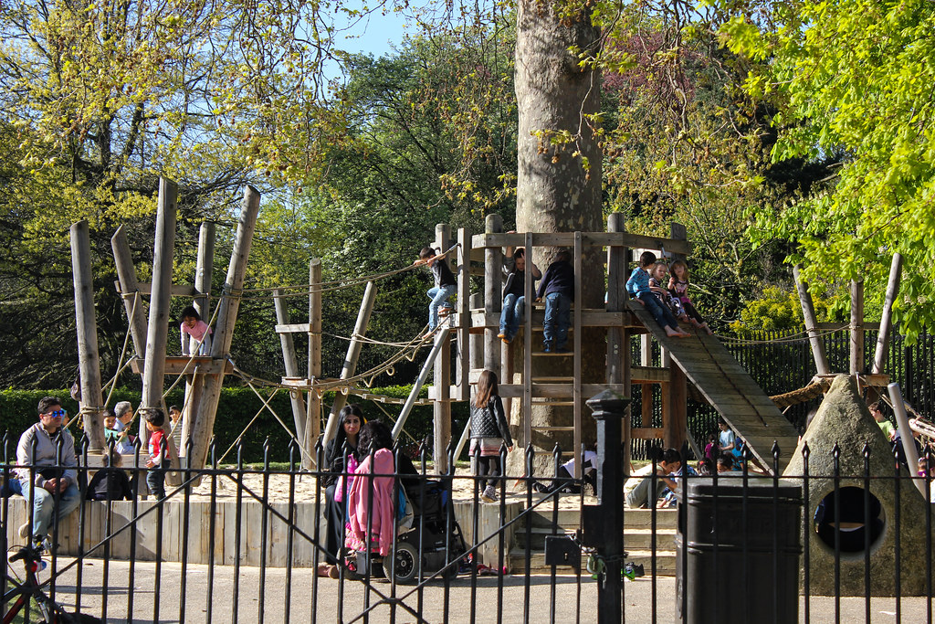 Niños jugando en un parque de juegos de madera en Regent's Park, con padres observando cerca.