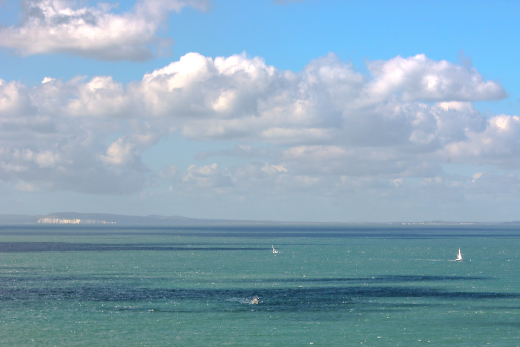 Vista panorámica del mar azul con veleros y acantilados blancos en la distancia bajo un cielo azul con nubes esponjosas.