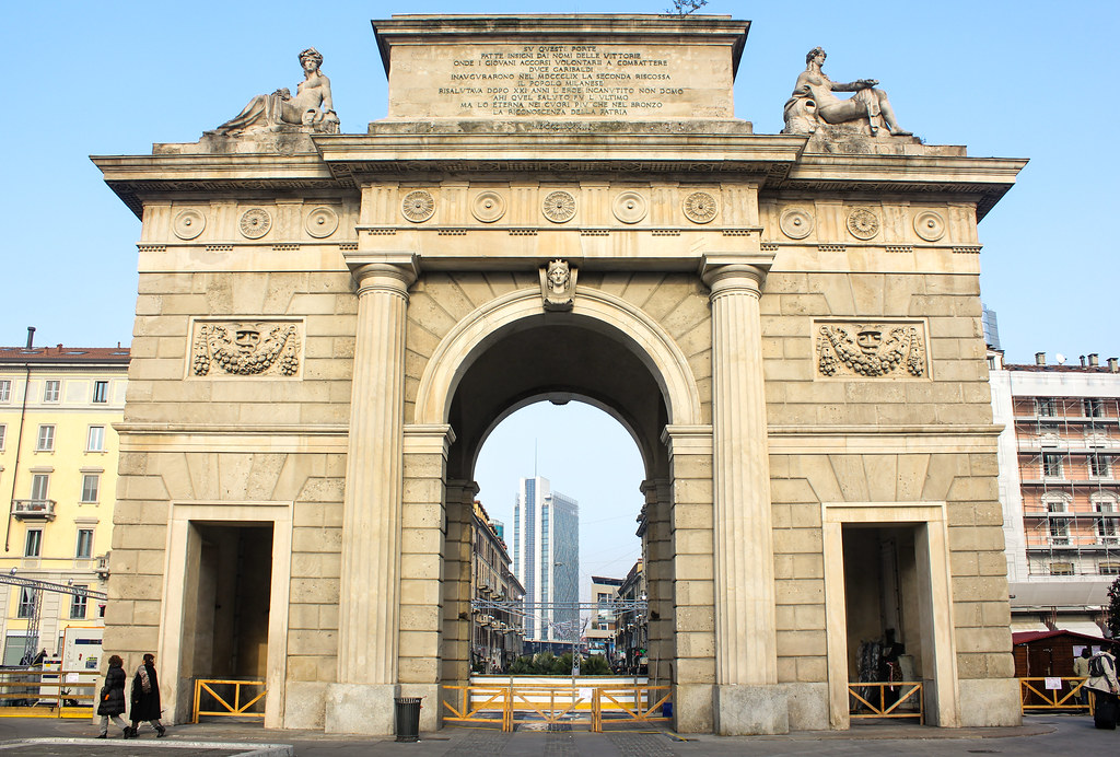 Puerta de la ciudad de Milán, Porta Nuova, con esculturas y peatones, día soleado.