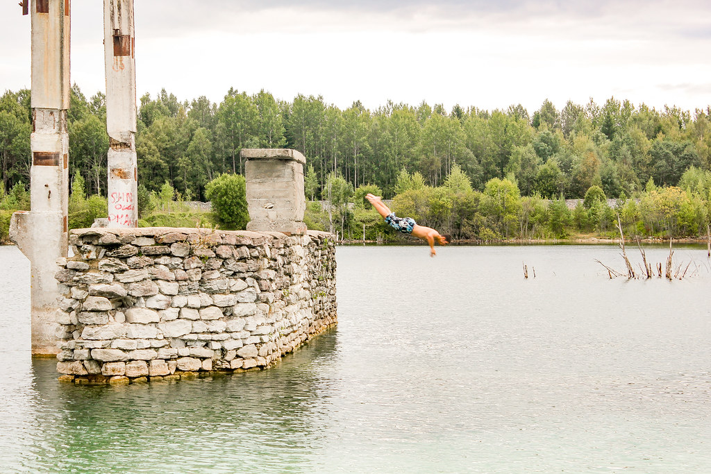 Coco saltando al agua desde una estructura de piedra en la cantera de Rummu, Estonia, con bosque al fondo.