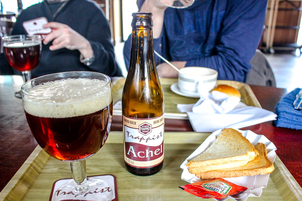 Cerveza trapense Achel servida en copa con botella y sándwich en mesa de cafetería.