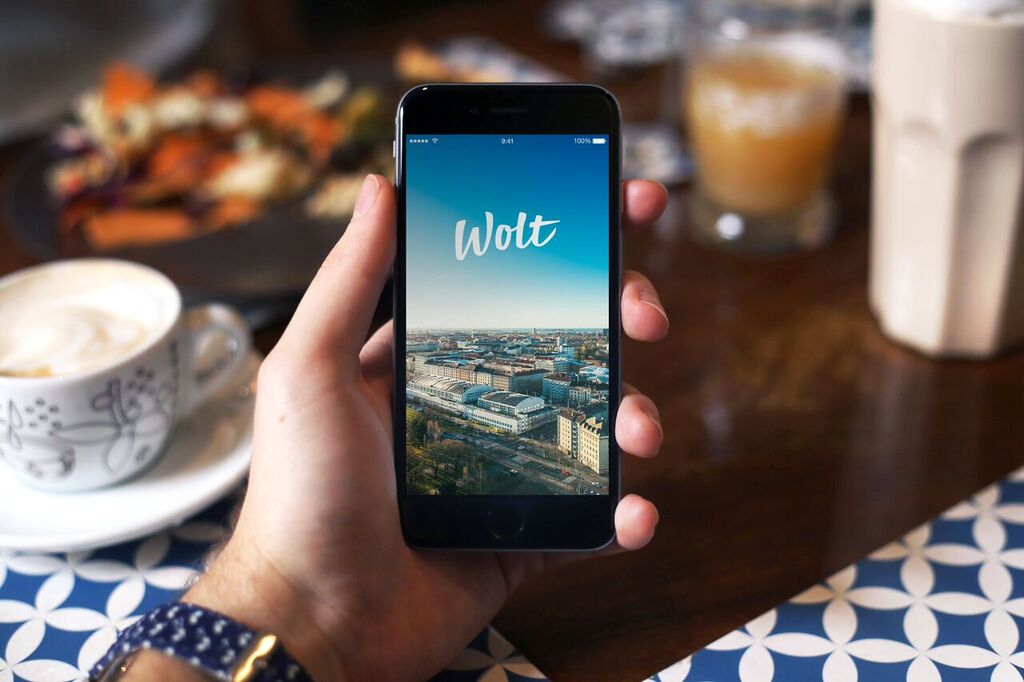 Mano sosteniendo un smartphone con la aplicación Wolt abierta mostrando vista aérea de la ciudad, con café y comida en la mesa.