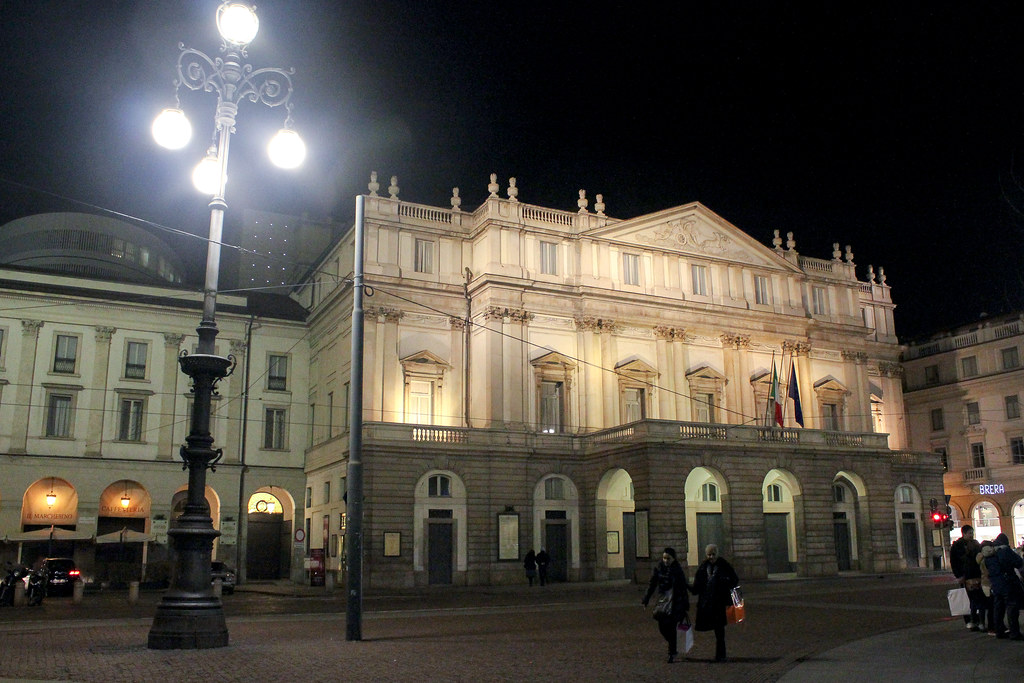Teatro alla Scala en Milán iluminado por la noche con peatones caminando en la plaza.