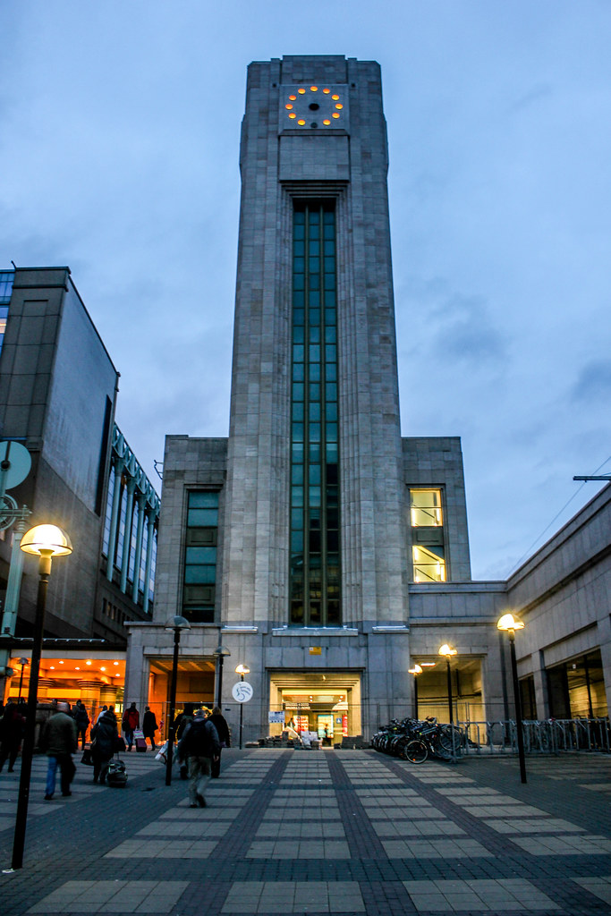 Torre de reloj art déco iluminada al atardecer en la estación de tren de Bruselas Nord, con personas caminando en la entrada.