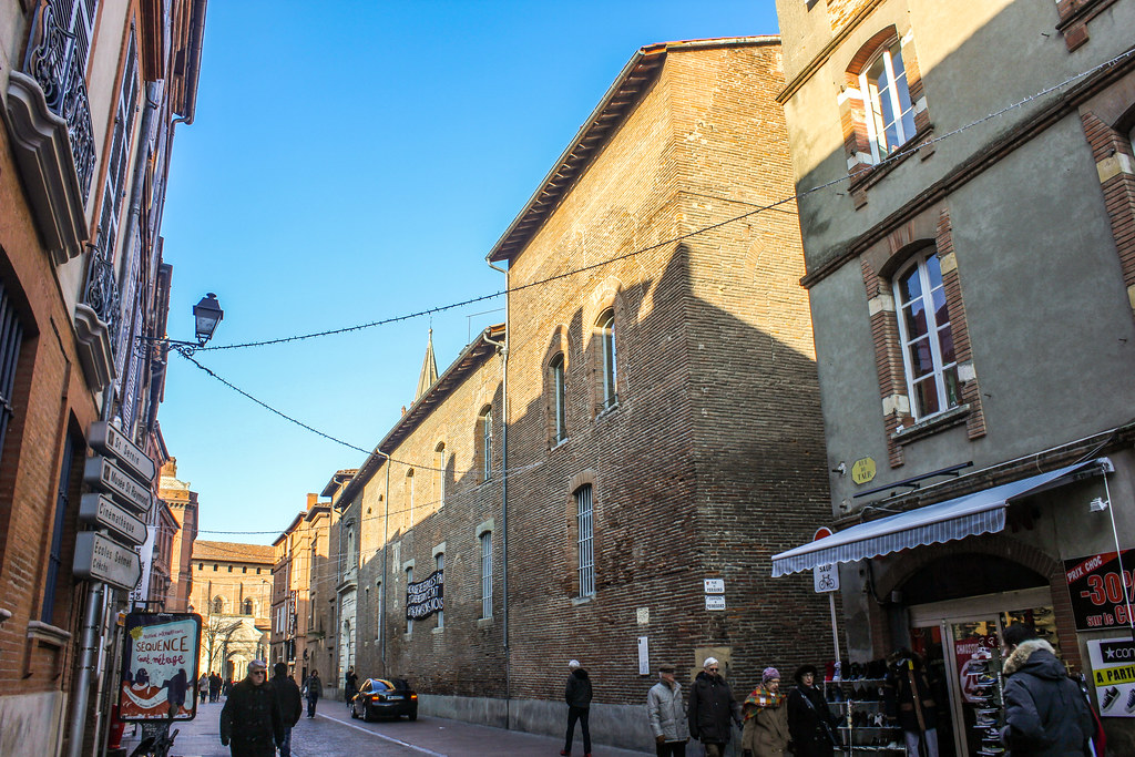 Transeúntes en una calle comercial de Toulouse con edificios de ladrillo y vistas al convento al fondo.