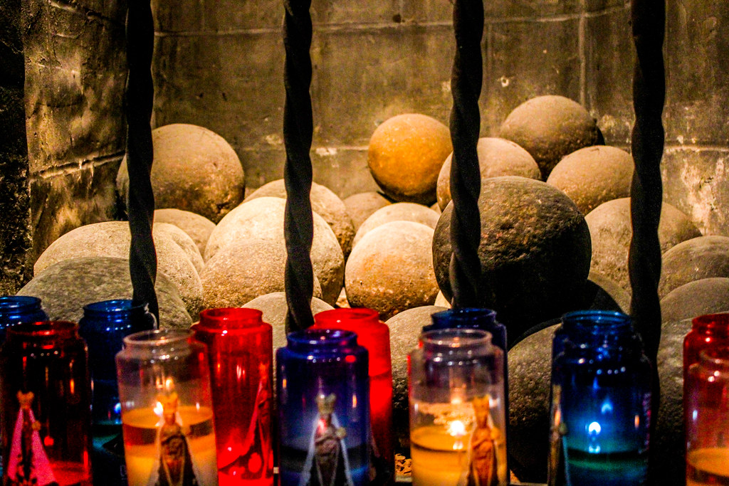 Velas de colores encendidas frente a una colección de antiguas bolas de cañón detrás de rejas de hierro en un ambiente oscuro y atmosférico.