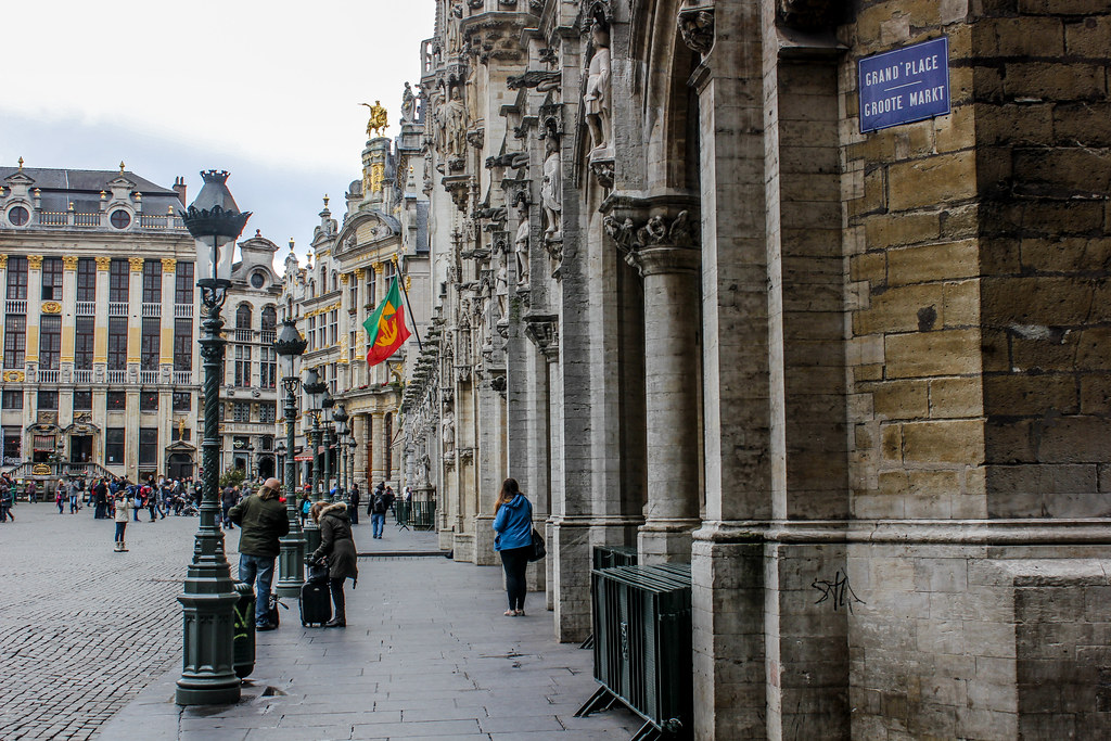 Vista de la Grand Place con la bandera de Bruselas y señalización bilingüe en la fachada de piedra del Ayuntamiento.