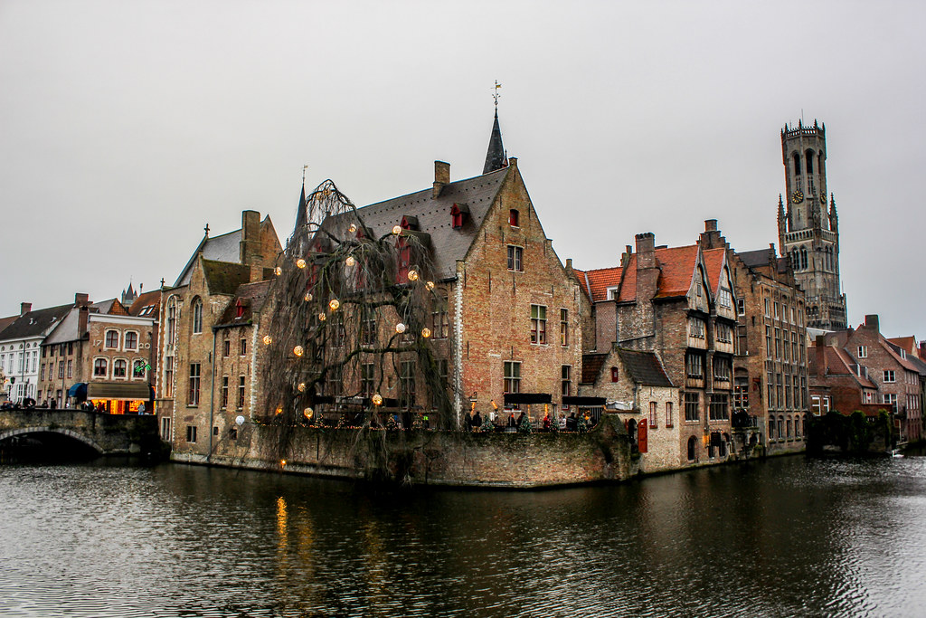 Vista pintoresca de casas históricas y el campanario de Belfort reflejados en el canal de Brujas, adornados con luces navideñas.